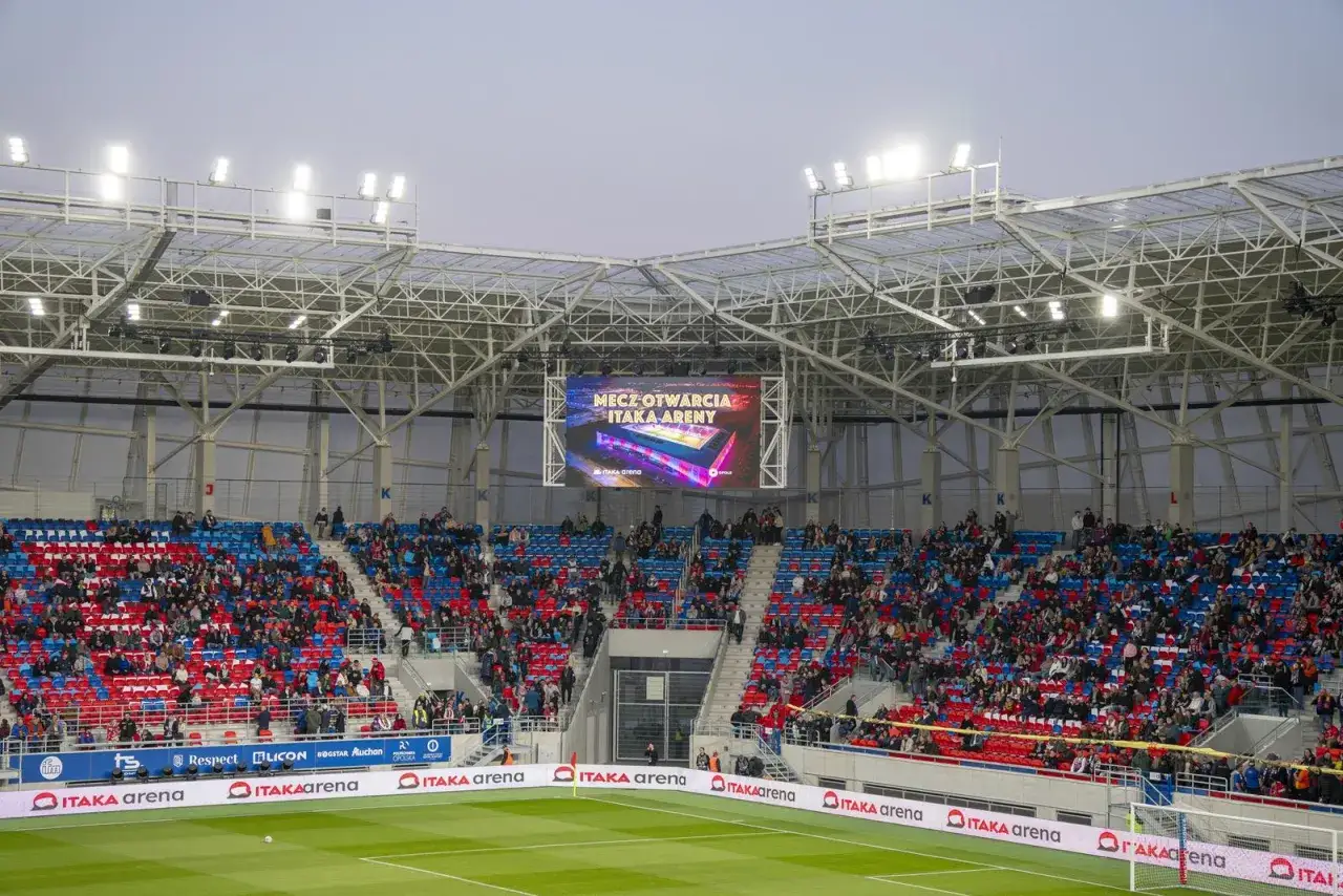 Widownia na stadionie Odra Opole podczas meczu otwarcia. Kolorowe trybuny wypełnione kibicami, nad nimi nowoczesny dach i telebim.
