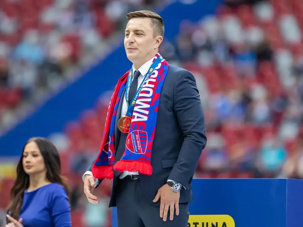 Michał Świerczewski w garniturze, z szalikiem Rakowa Częstochowa i medalem na szyi, symbolizuje sukcesy i majątek klubu.