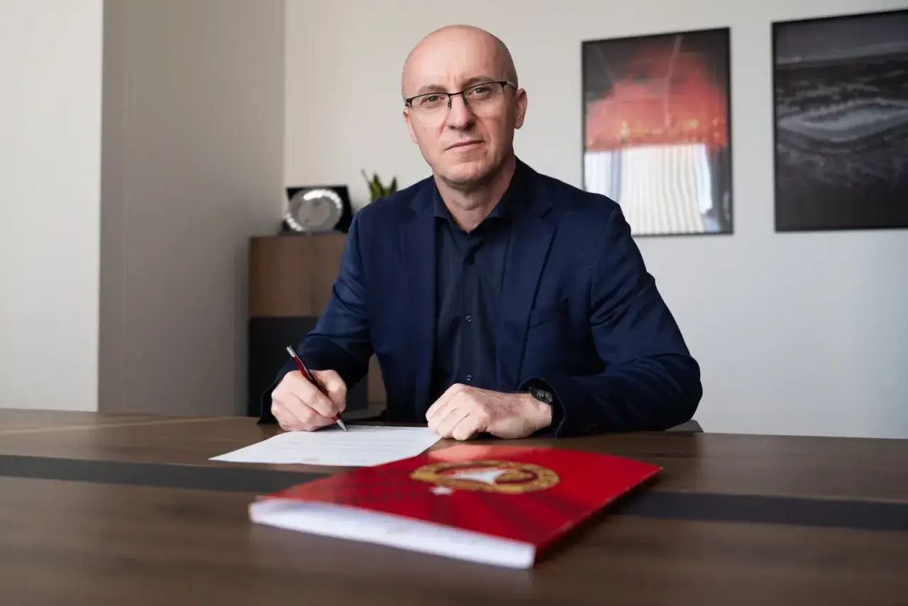 Nowy trener Widzewa podpisuje kontrakt. Mężczyzna w okularach i granatowej marynarce siedzi przy biurku, pisząc piórem.