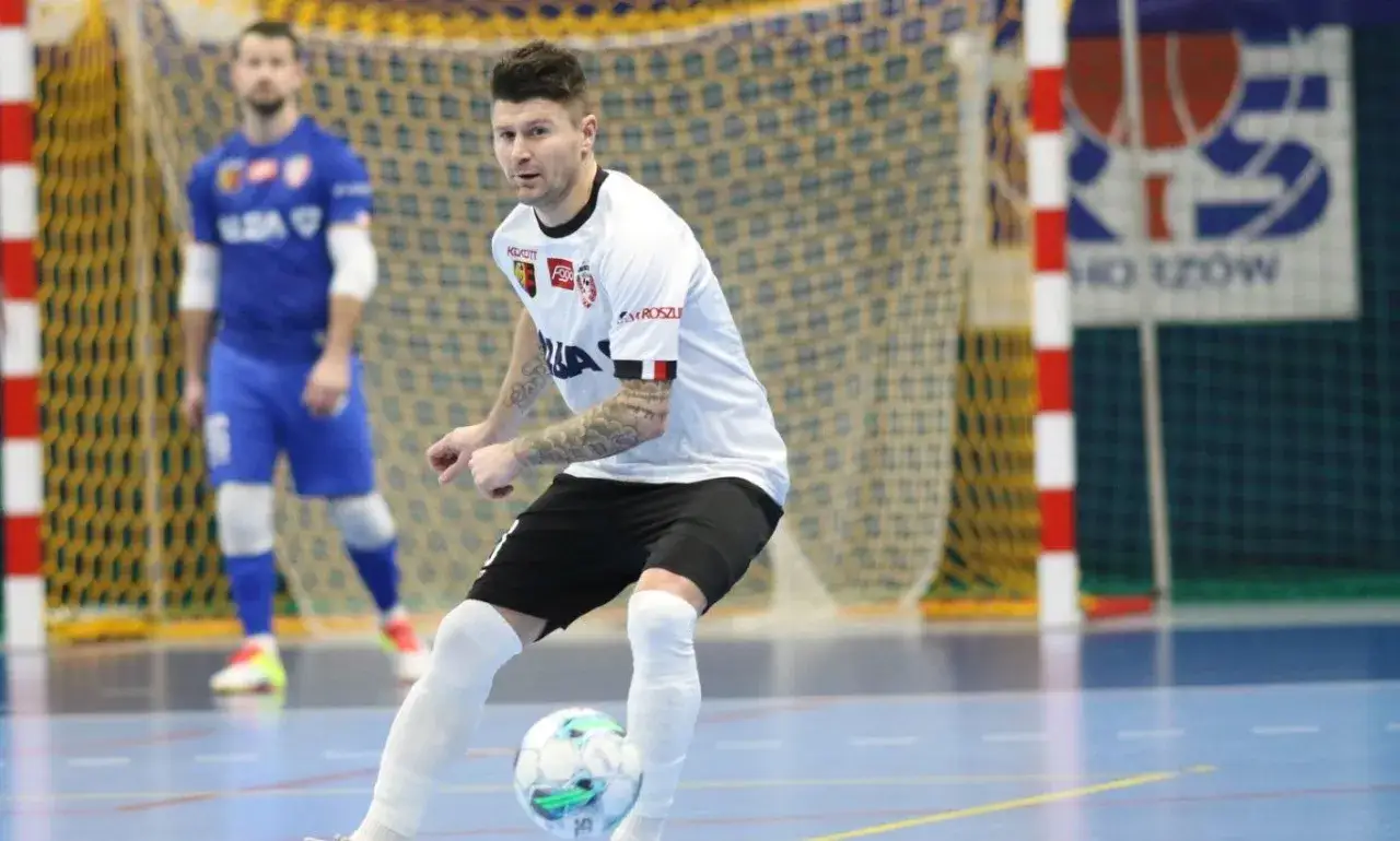 Piłkarz w białej koszulce drybluje piłką na hali. To właśnie futsal – szybka gra, która wciąga!
