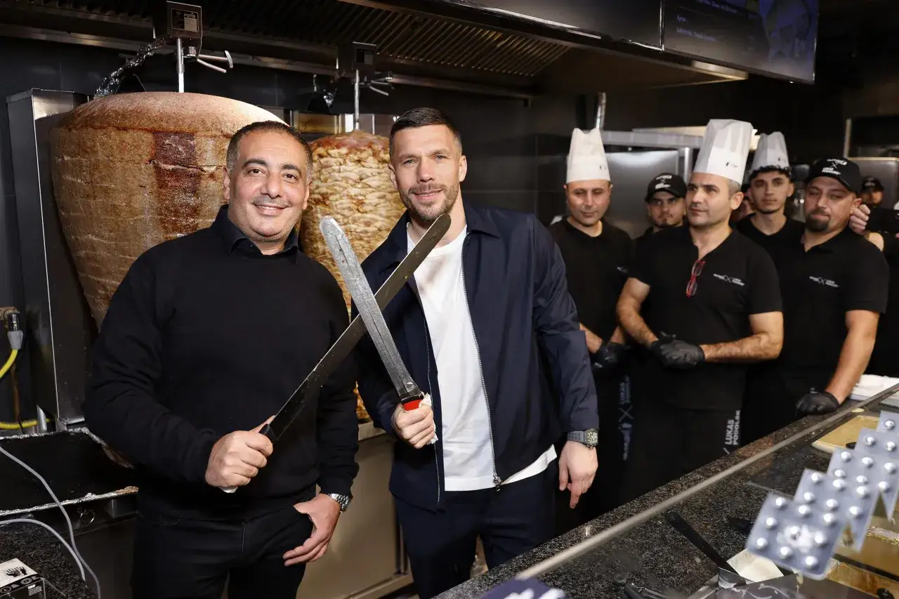 Dwóch mężczyzn z nożami w rękach pozuje przed kebabem. Jeden z nich to Lukas Podolski, który inwestuje w biznes, budując swój majątek.