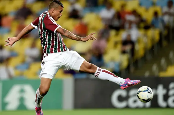 Piłkarz w koszulce Fluminense kopie piłkę na tle trybun.